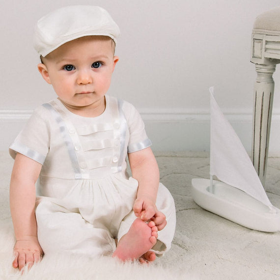 Baby boy sitting down and wearing the Owen Linen Romper featuring silk ribbon in ivory and light blue across the front bodice and sleeves