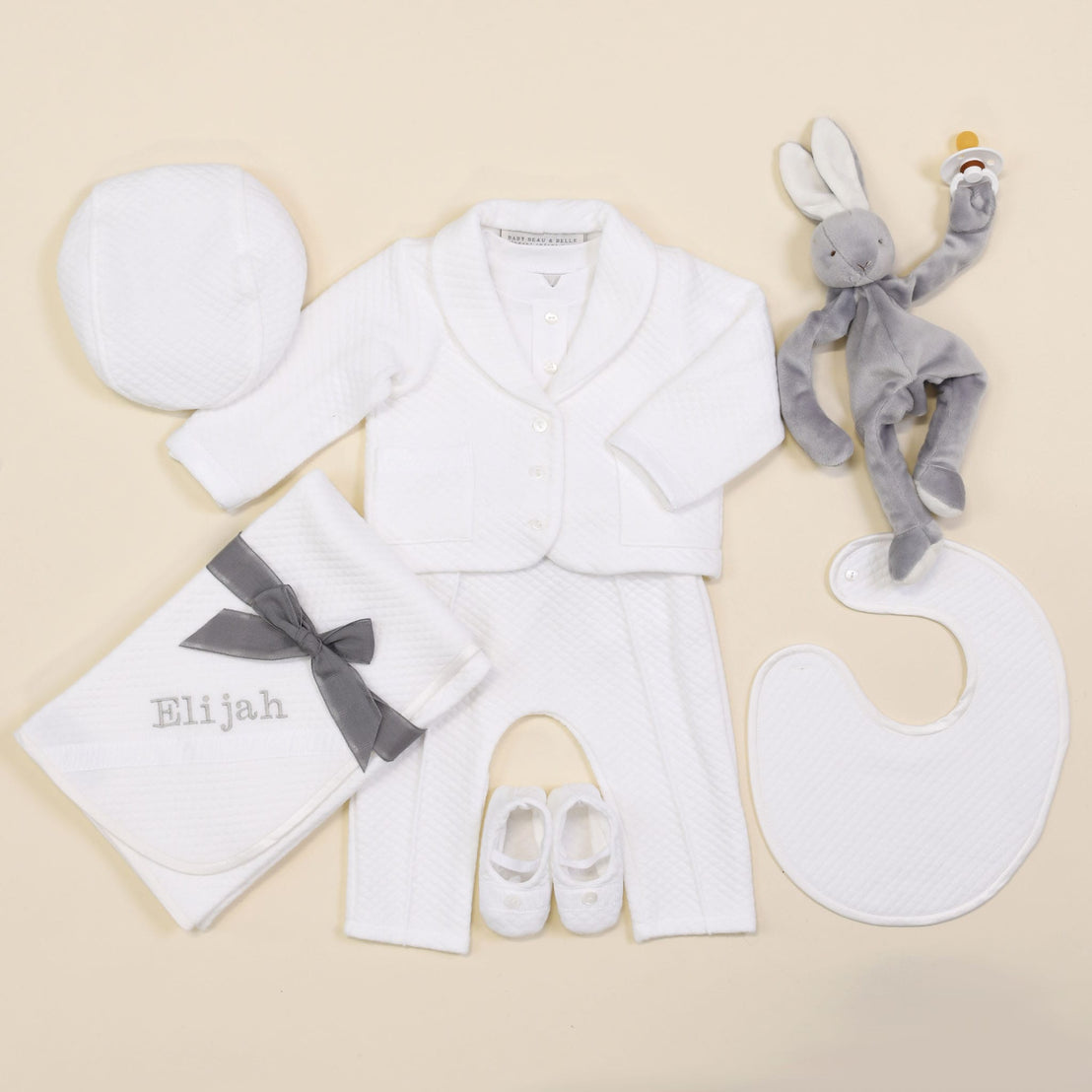 The Elijah Newborn 3-Piece Suit features a white coming home outfit set with a jacket, pants, hat, bib, and booties. Accompanying this charming ensemble is a personalized blanket labeled "Elijah" and a gray rabbit plush toy that holds an orange rattle. Everything is beautifully arranged on a soft beige background.
