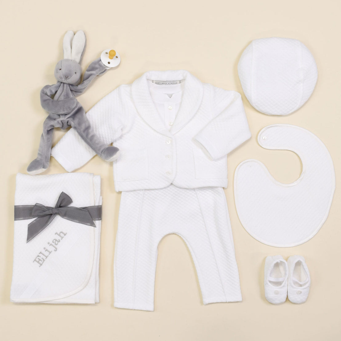 A flat lay display of baby essentials: a gray stuffed rabbit rattle, the Elijah Newborn 3-Piece Suit featuring a white jacket, pants, and hat as a coming home outfit, complemented by a matching bib and booties, along with a personalized white blanket embroidered with the name "Elijah.