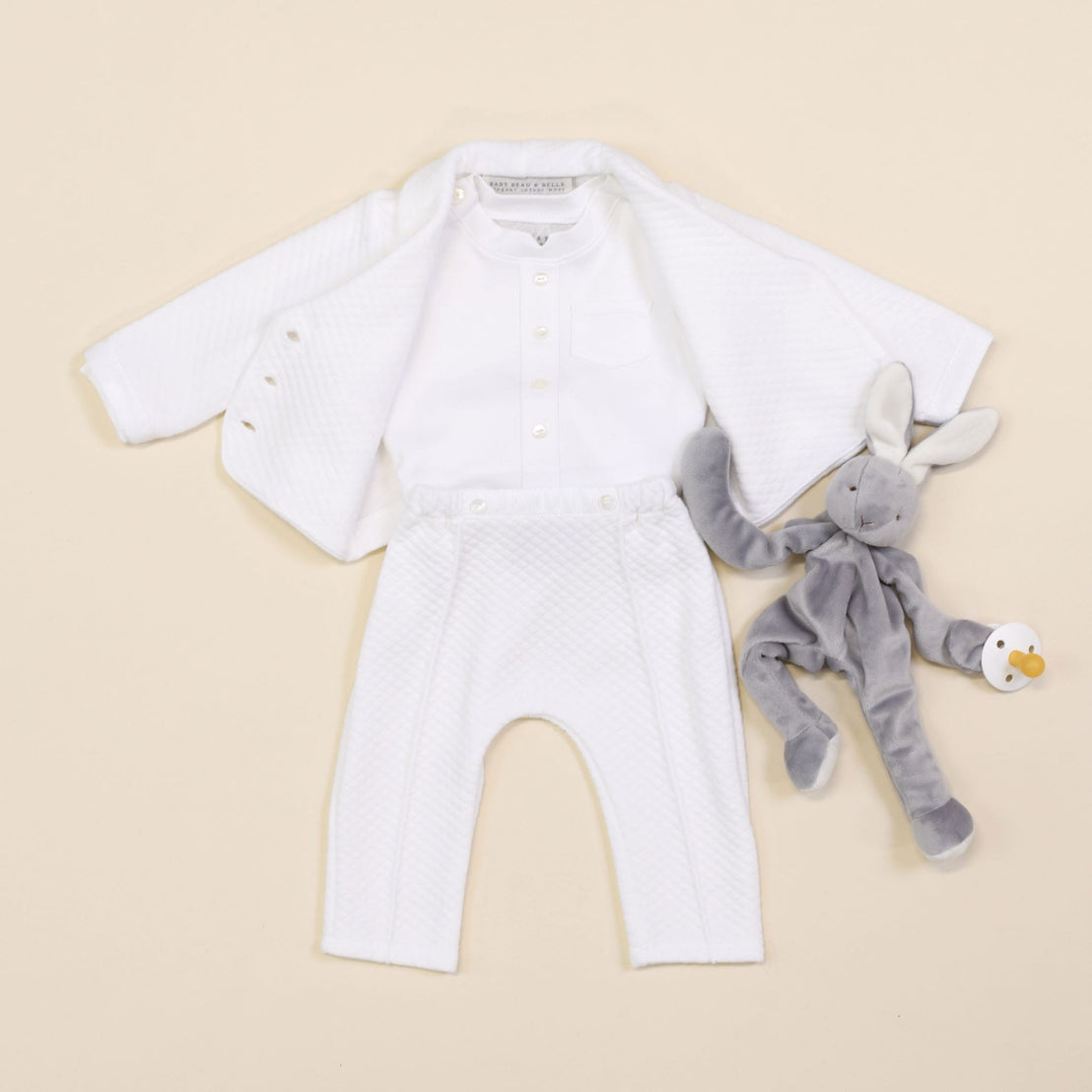 The Elijah Newborn 3-Piece Suit, which includes a white quilted jacket, button-up shirt, and pants, is laid flat. Next to it, a gray stuffed bunny holding a pacifier rests on a light beige background, making it an ideal choice for a baptism outfit.