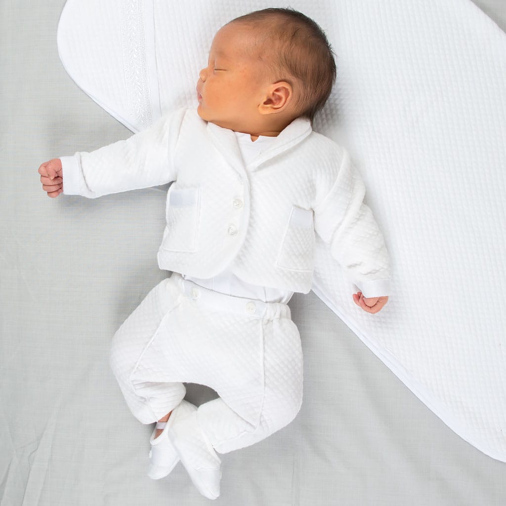 A sleeping baby wearing the Elijah Newborn 3-Piece Suit, featuring a white quilted design, lies peacefully on a light gray surface with a coordinating white quilted blanket beside them. One arm is gently stretched out as they rest.