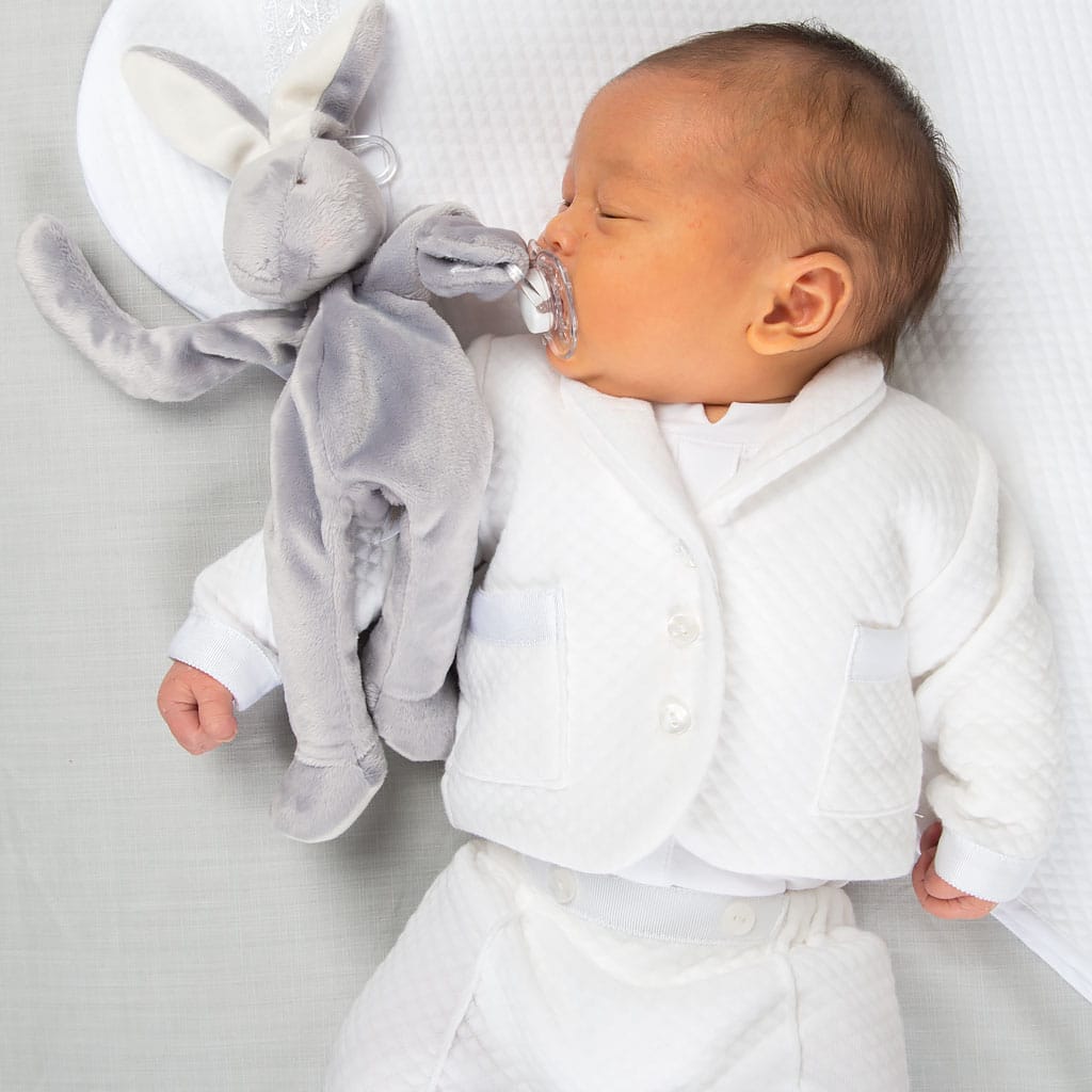 A sleeping baby, dressed in the Elijah Newborn 3-Piece Suit, cuddles a gray plush bunny while holding a pacifier in its mouth. The little one is lying on a soft, white surface.