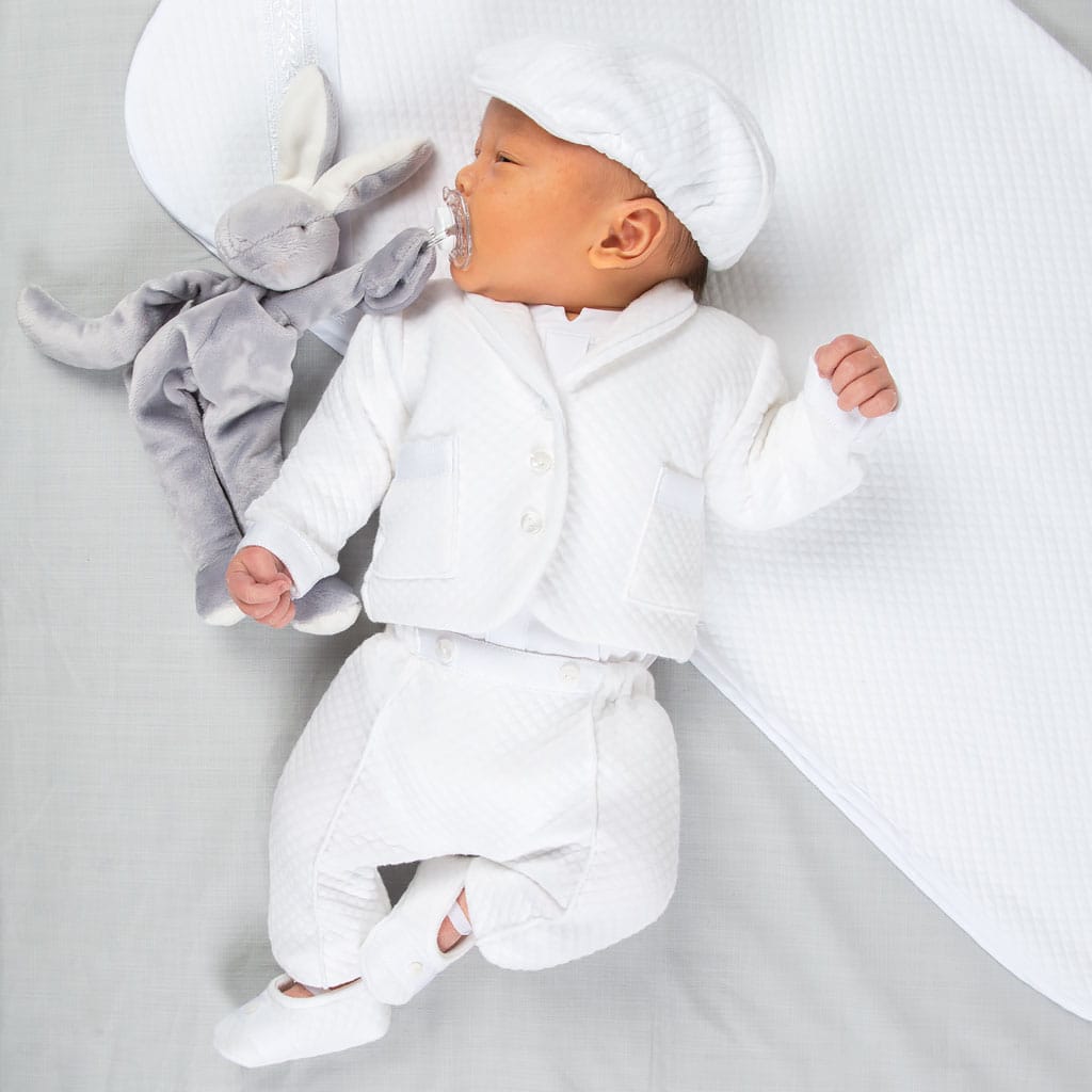 A baby wearing the Elijah Newborn 3-Piece Suit and matching cap lies on a soft, white surface next to a gray stuffed rabbit, serenely holding a pacifier in their mouth.