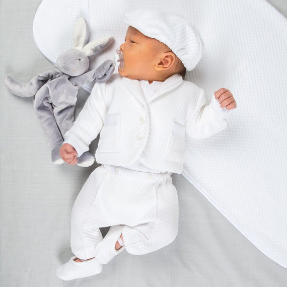 A baby wearing the Elijah Newborn 3-Piece Suit and matching cap lies on a soft, white surface next to a gray stuffed rabbit, serenely holding a pacifier in their mouth.
