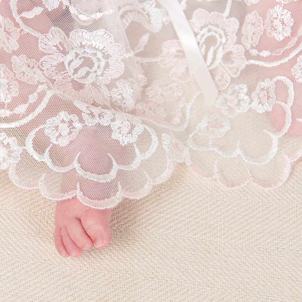 A close-up image of a baby's foot peeking out from underneath a Joli Christening Gown with floral patterns on a neutral woven background.