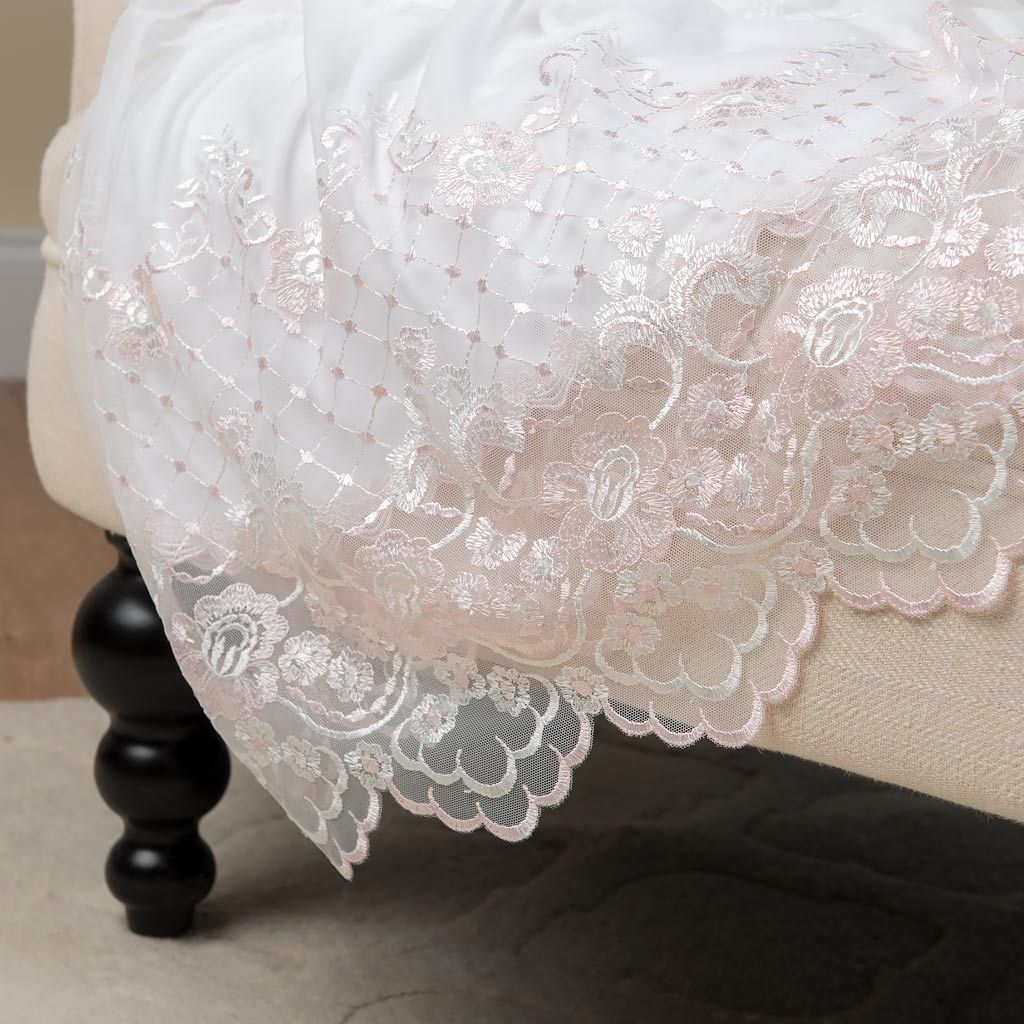 Close-up of an elegant lace-edged Joli Christening Gown draped over a bed, highlighting detailed floral embroidery and scalloped edges against a neutral-toned bedroom background.