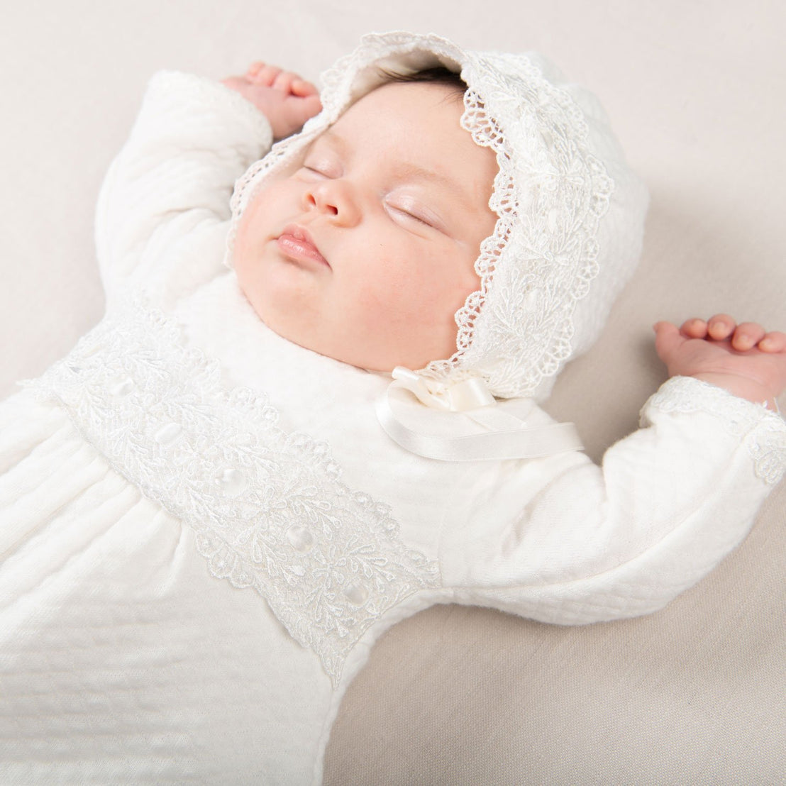A newborn baby sleeps peacefully, dressed in a Madeline Quilted Newborn Romper and matching bonnet from a boutique, with arms gently rested to the sides on a soft beige background.