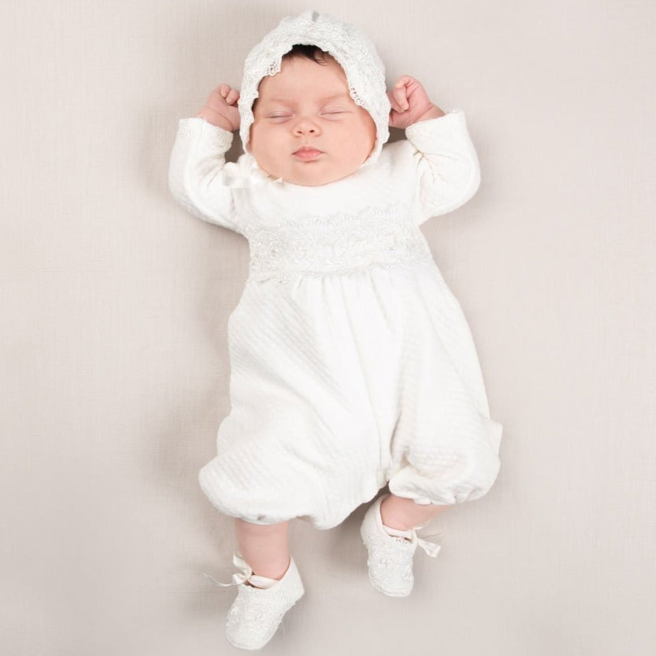 A newborn baby wearing a White Madeline Quilted Newborn Romper outfit and hat, lying on a soft beige background with arms stretched out.